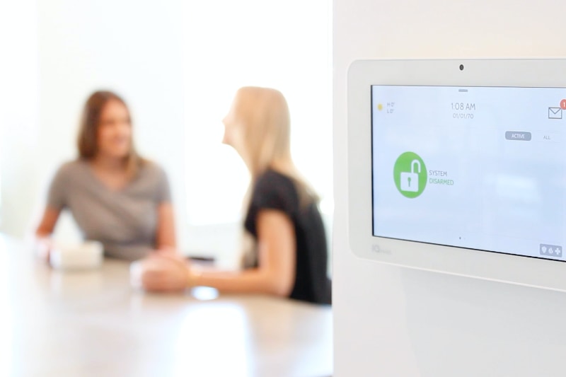 two women talking at kitchen table with security smart home panel on wall